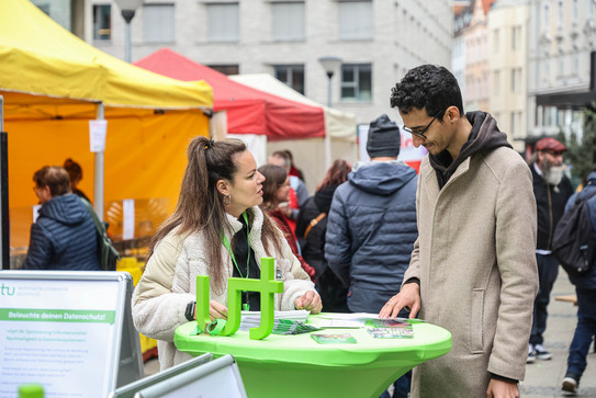 Photo: At a city festival in Dortmund city center, a woman and a man stand at a table and talk. In the background you can see colorful stalls. The TU Dortmund University logo is on the table.